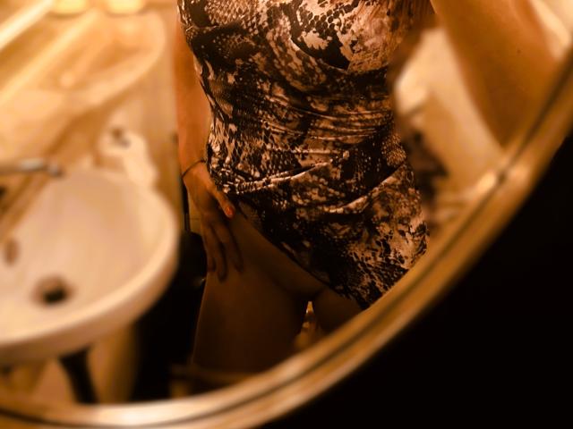 Una mujer delgada, con un vestido ajustado y estampado, lo levanta hacia un lado, revelando un poco de su vello púbico blanco y cremoso en un reflejo del espejo..