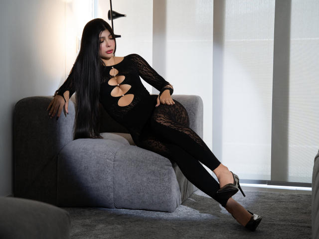 NatyJanssen with long black hair, wearing a black lace bodysuit with cutouts, sitting on a gray chair, legs crossed, black high heels, and a relaxed, confident expression.