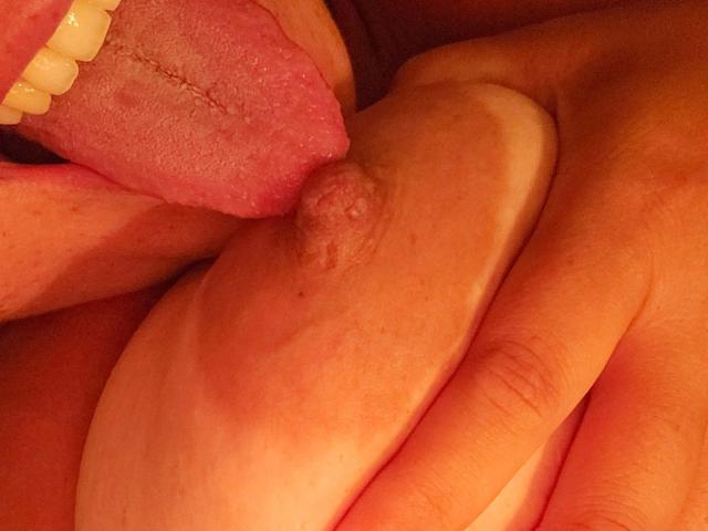 LadyChina-hot's tongue is licking her titty, with her finger gently holding the areola. Her pink tongue and white teeth are visible. The skin is slightly freckled.