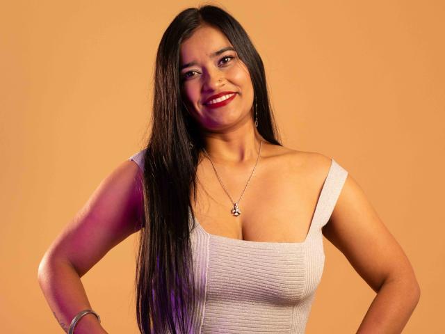 ValeriaHotVelez with long black hair, smiling, red lipstick, white ribbed tank top, small silver necklace, hands on hips, orange background, medium breasts, confident, cute, attractive.