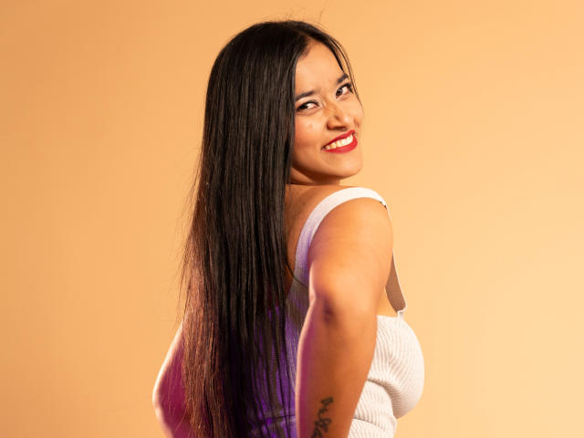 ValeriaHotVelez has long black hair, red lipstick, and a white tank top, smiling with her back to the camera, showing her ass and tattoo on her right arm.