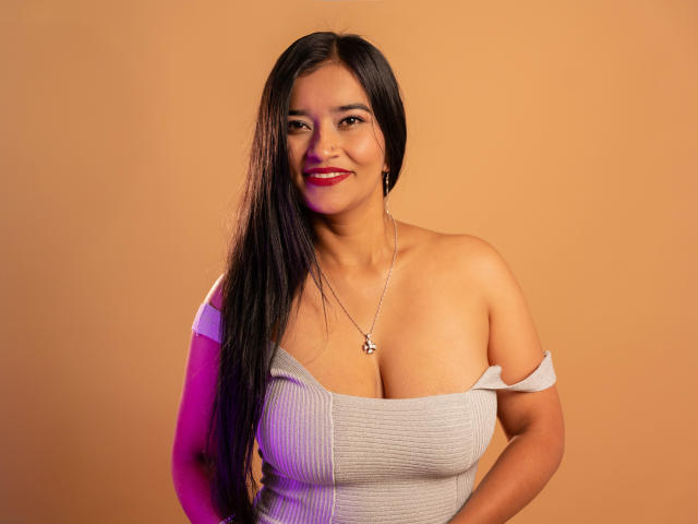 ValeriaHotVelez, brown-skinned woman with long black hair, smiling, wearing a gray off-shoulder top, revealing her large tits, silver necklace, pinkish-purple light on her right.