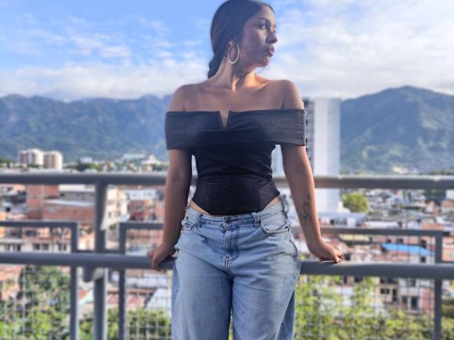 HelenaJones with bare tits in an off-shoulder black top, blue jeans, and hoop earrings, standing on a balcony, looking to the side, mountains and city in the background.