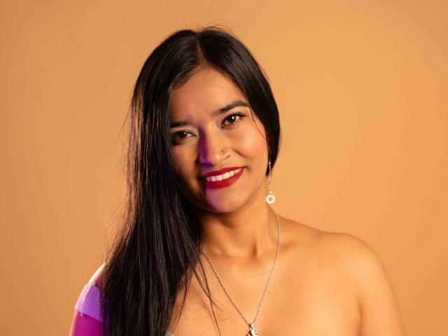 ValeriaHotVelez has long black hair, red lipstick, and a nose ring, smiling with exposed boobs, wearing a necklace, against a warm orange background.