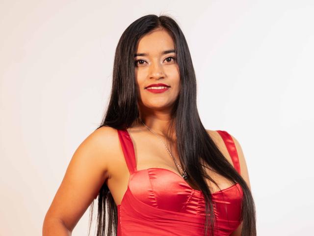 ValeriaHotVelez with long black hair, red satin dress, showing her tits, smiling with red lipstick, standing against a white background, looking cute and sexy.