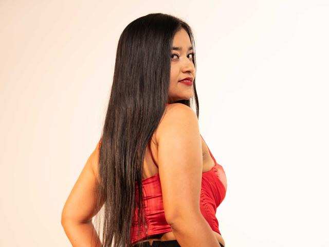 ValeriaHotVelez with long black hair, red lace-up top, looking over her shoulder, showing her back and small, perky tits, standing against a plain white background.