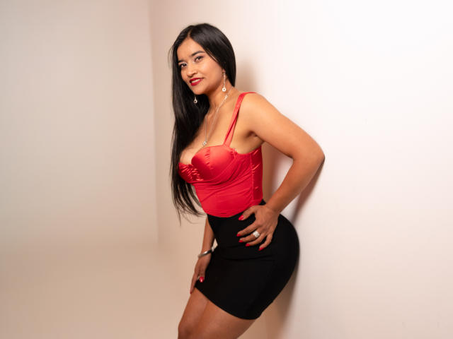 ValeriaHotVelez with long black hair, red satin top, showing big boobs, black skirt, red nails, hand on hip, standing against white wall, looking at camera with red lipstick.