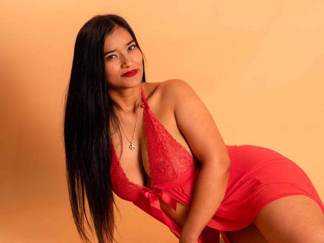 ValeriaHotVelez with long black hair, red lingerie, and a silver necklace, leaning forward, showing her tits, with a slight smile and red lipstick, against a warm orange background.