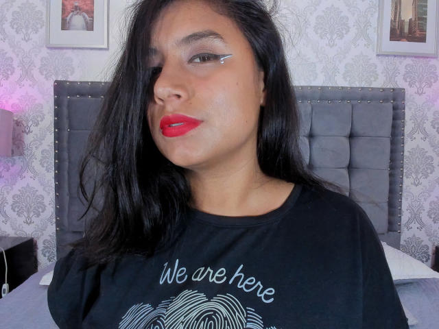 AbrilMoon with dark hair, red lipstick, and black eyeliner, wearing a black shirt, showing her tits, sitting in front of a grey upholstered headboard.