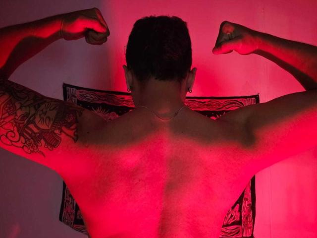 ConnieXMarc, torse nu, les bras tendus, une lumière rouge sur le dos, un tatouage sur le bras droit, cheveux courts et foncés, un collier, musclé, dos à la caméra, mettant en valeur une carrure forte..