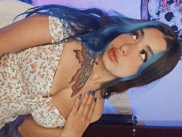 ConnieXMarc avec des mèches bleues, portant une robe fleurie, montrant un tatouage de papillon sur sa poitrine, regardant vers le haut, ongles peints en bleu, sexy, mignonne, grande poitrine, lumière douce..