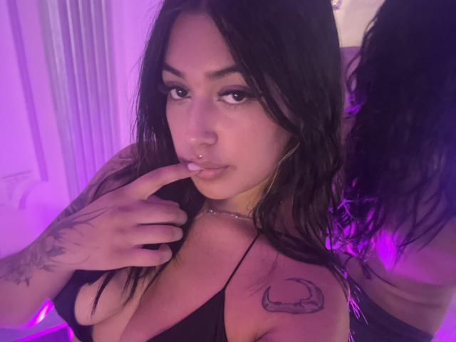 KatianPierce, com cabelos pretos longos, piercing no nariz e tatuagens no ombro e no pulso, mordendo o dedo, vestindo uma blusa preta, com iluminação roxa. Seios visíveis na blusa com decote baixo..