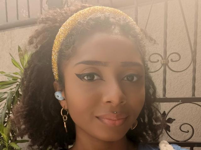 MeganWlliams with curly brown hair, gold glitter headband, black eyeliner, blue plug earrings, gold hoop earrings, nose ring, smiling, light brown skin, and green plant in the background.