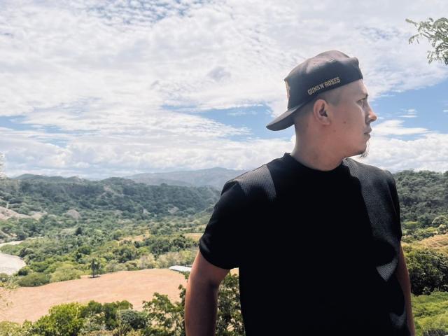 LianaAndAsaff, a chubby dude with a black cap, black shirt, and short hair, stands with his back to the camera, looking off to the side. Bright, cloudy sky.