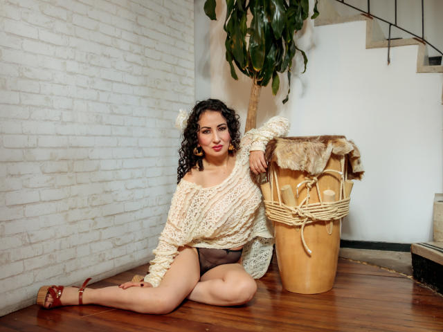 SamanthaAntolin with curly dark hair, white knit sweater, brown shorts, red heels, sitting on wooden floor, next to wicker basket with fur, smiling, hand on basket.