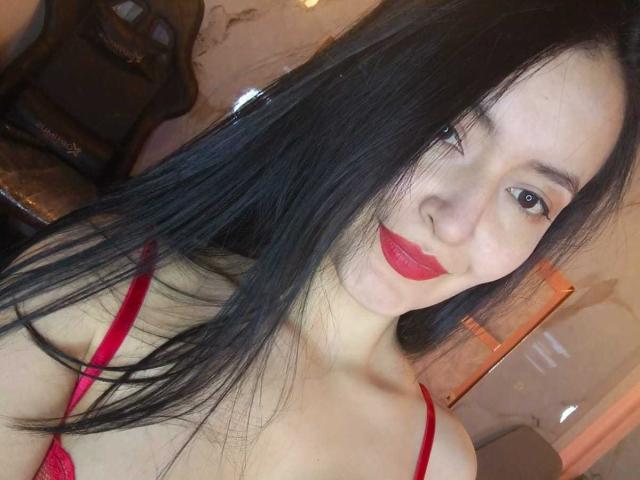 AriaBonnet with long black hair, red lipstick, and red bra, showing her tits, smiling at the camera, in a dimly lit room.