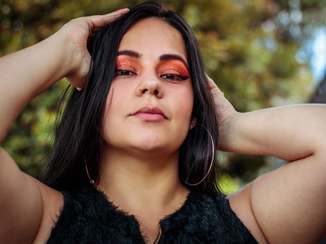NohayMuller has dark hair, orange eye makeup, and hoop earrings, with hands in hair, wearing a black furry top, looking confident. Her tits are slightly visible.