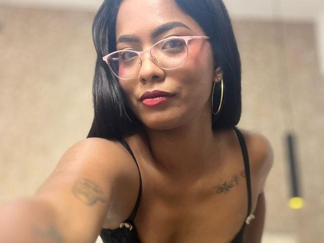 ChloeBrownOne with black hair, pink glasses, hoop earrings, red lipstick, and tattoos on her shoulder and chest, wearing a black spaghetti strap top, showing her tits.