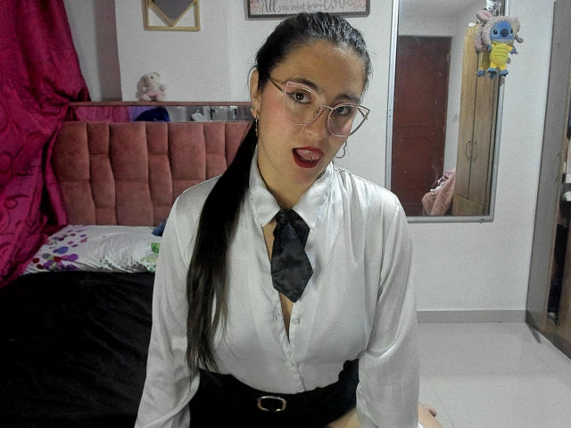VenusSweetPalmer with long black ponytail, wearing glasses, white shirt with black tie, black waistband, sitting, red lips, looking at camera, bedroom, pink headboard, stuffed toy in mirror.