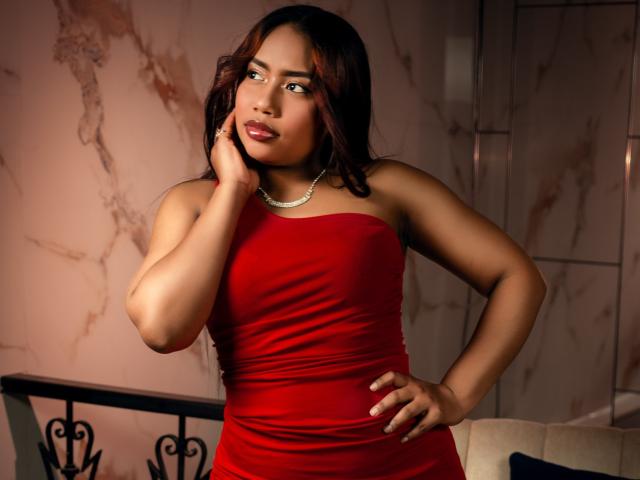 MiaXgate with dark red dress, necklace, hand on cheek, and hand on hip, looking to the side, showing her tits and ass, in a dimly lit room.