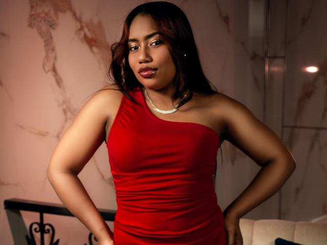 MiaXgate with dark brown hair, wearing a red one-shoulder dress, hands on hips, looking slightly to the side, wearing a silver necklace, medium brown skin, full lips, small tits.