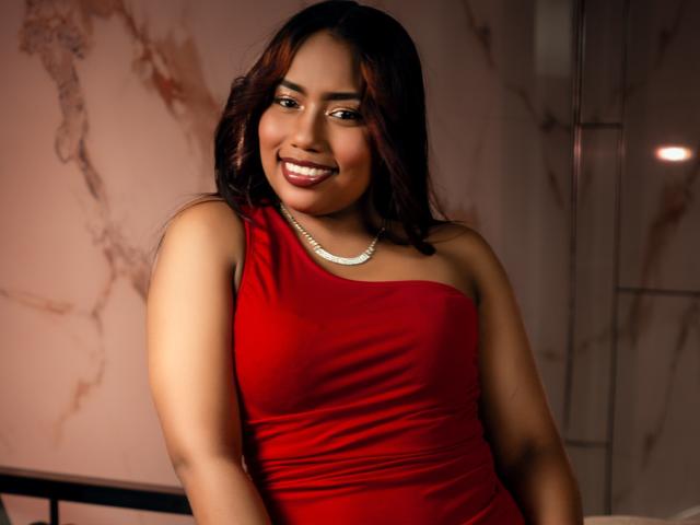 MiaXgate with dark red hair, wearing a red dress and white pearl necklace, smiling at the camera, showing her tits slightly, in a warm-lit room.