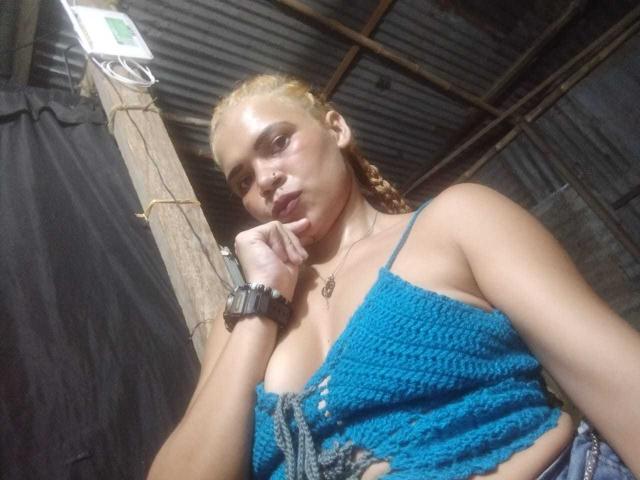 DomiEvans, com tranças loiras, vestindo uma blusa azul de crochê que expõe seus seios, usa um relógio preto e um colar, e posa com o dedo nos lábios..