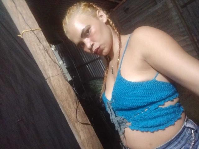 DomiEvans with blonde braids, wearing a blue, crocheted, sleeveless top that shows her small tits and a cut-out below her breasts, standing in a dimly lit room.