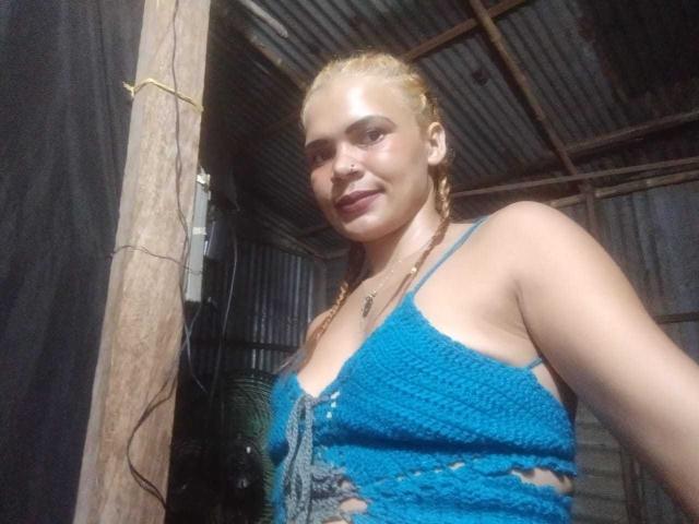 DomiEvans, avec des tresses blondes, portant un haut bleu en crochet avec des découpes sur les côtés, montrant ses seins, et un petit anneau au nez, souriant dans une cabane rustique éclairée par le soleil, avec un toit en métal..