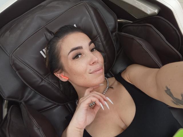 ConnyShellie is lying in a brown leather chair, wearing a black tank top, showing her tits, and smiling with a lip piercing, long nails, and a tattoo on her arm.