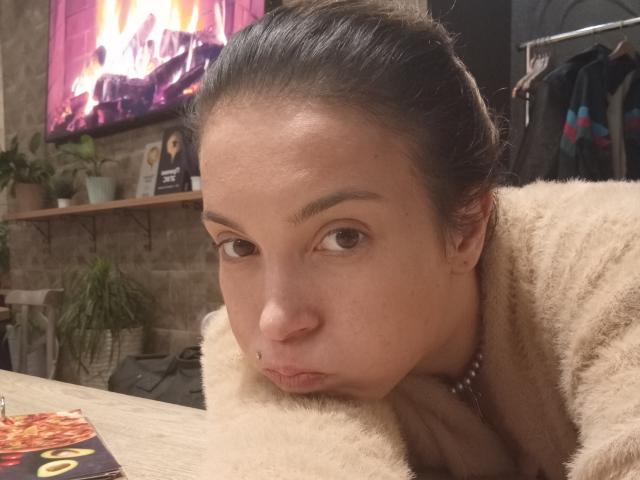 ConnyShellie with pouty lips, brown eyes, and dark hair in a bun, wearing a fluffy beige coat, and a pearl necklace, leaning on a wooden table.