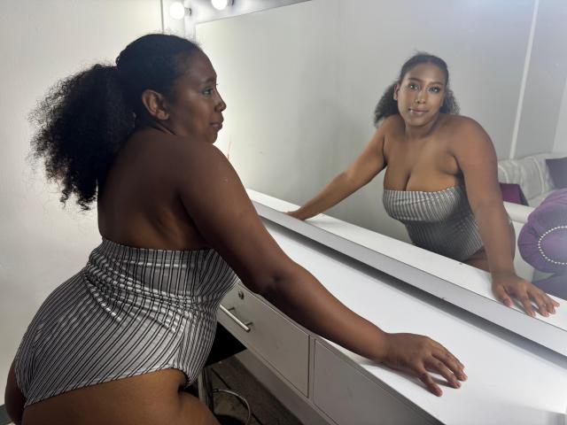 RebecaSolis, curvy with dark skin and curly hair in a ponytail, wears a striped strapless one-piece. She leans on a vanity, staring at her reflection in the mirror.