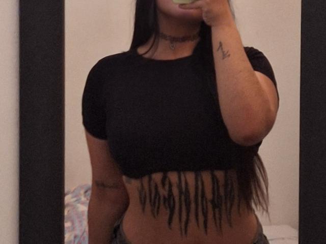 DesireSweet, com longos cabelos pretos, uma cropped top preta que mostra seus seios, e uma tatuagem na parte inferior do abdômen com linhas pretas penduradas, segurando um celular em um selfie no espelho..