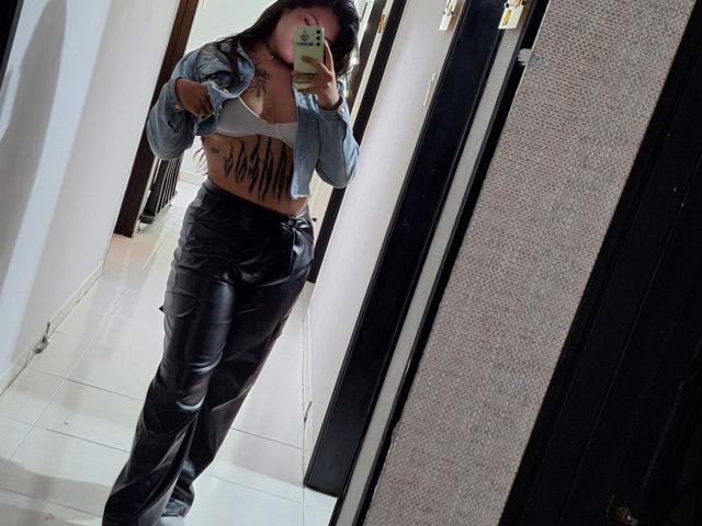 DesireSweet vestindo calças de couro pretas, jaqueta cinza e sutiã branco, mostrando uma tatuagem em sua barriga lisa e bronzeada, tirando uma selfie no espelho em uma loja, com o celular na mão..