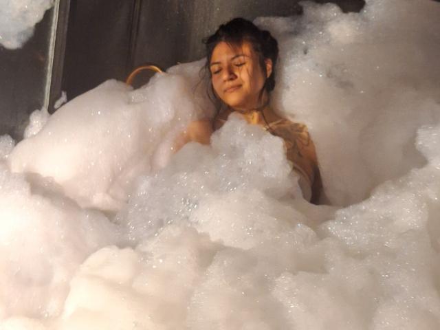 DesireSweet with her tits partially covered in thick white bubbles, eyes closed, and a calm expression, sitting in a foamy bathtub. She has a gold necklace on.