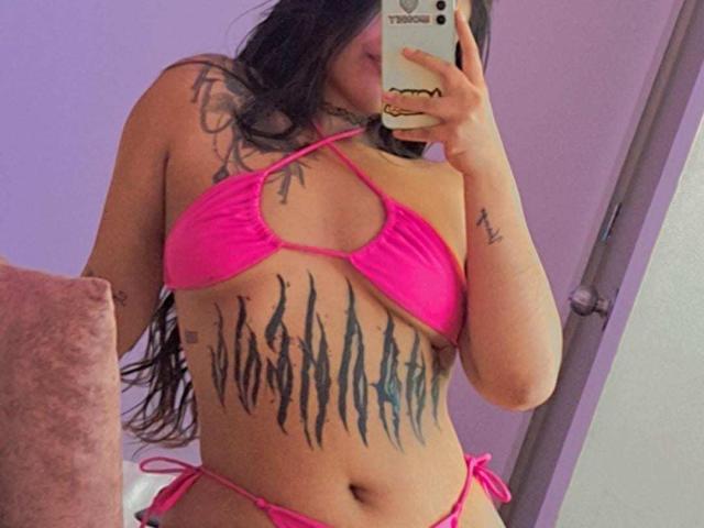DesireSweet, con lunghi capelli neri, un bikini rosa e grandi tatuaggi neri sul torso, sta scattando un selfie allo specchio, mostrando i suoi seni e i suoi glutei, e tiene in mano un telefono con una custodia..