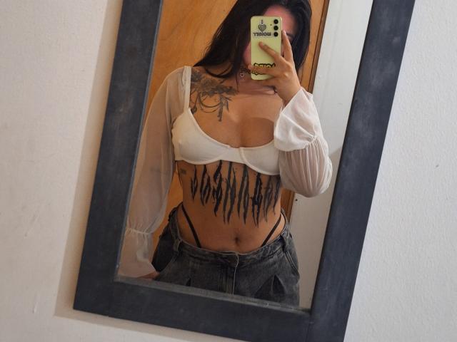 DesireSweet mit mittelgroßer Brust, in einem weißen BH und mit durchsichtigen Ärmeln, mit schwarzen Tätowierungen auf der Brust, hält ein Telefon vor einem Spiegel, trägt dunkle Jeans und hat langes, dunkles Haar..