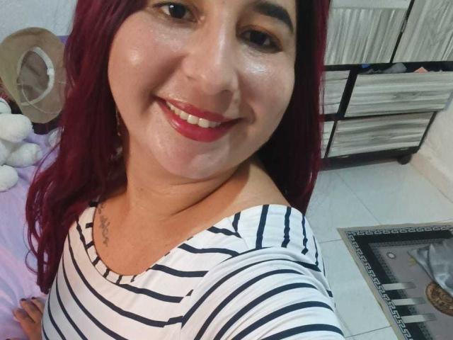 BerenicePretty with red hair, smiling, wearing a striped shirt, showing her tits slightly with a low neckline, tattoo on left chest, red lipstick, white tile floor, and a mirror.