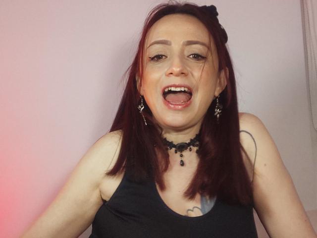 CatalinaMonyque with red hair, black tank top, black choker, earrings, and tongue out, smiling. Boobs slightly visible under tank top. Tattoo on right chest. Pearly white teeth.