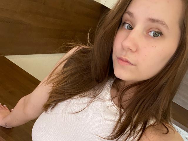 HotCaramells with light skin, brown eyes, and long brown hair, wearing a white sleeveless top, showing her tits and slightly puffy ass, leaning on a wooden surface.