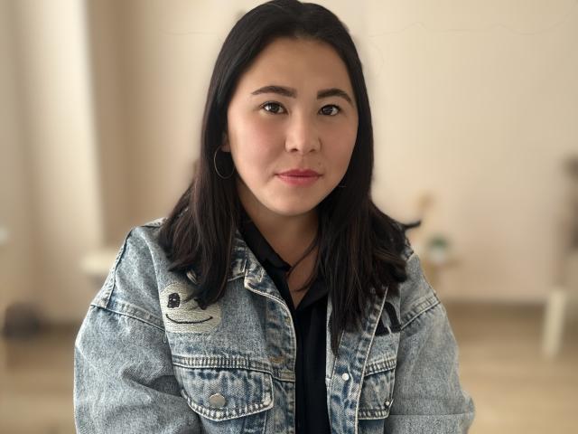 MonicaGeller with medium brown skin, black hair, wearing a denim jacket and black shirt, small hoop earrings, slight smile, soft light, focused face, casual style.