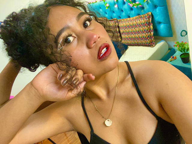 SophieSonyas with curly black hair, dark red lipstick, gold necklace, black top, looking up, hand on cheek, brown eyes, medium brown skin, small gold pendant, relaxed expression.