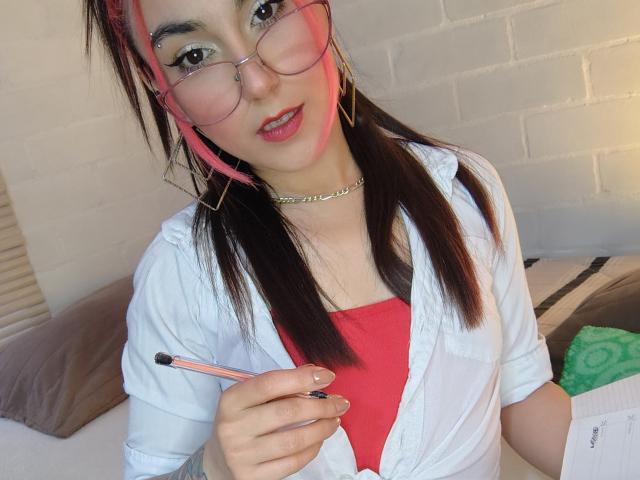 AmyStreetwise69 with pink-tinted glasses, pink streaks in brown hair, white shirt, red top, holding pencil, choker, looking at camera, bed in background, slightly smiling, pink lipstick.