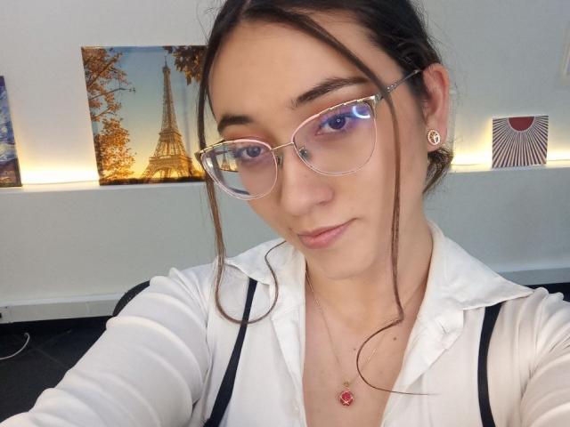 SwettieSammy with glasses, white shirt, and red necklace, has blue eyes, brown hair, and a cute face, showing a subtle smile. Her tits are small, and she's looking at the camera.