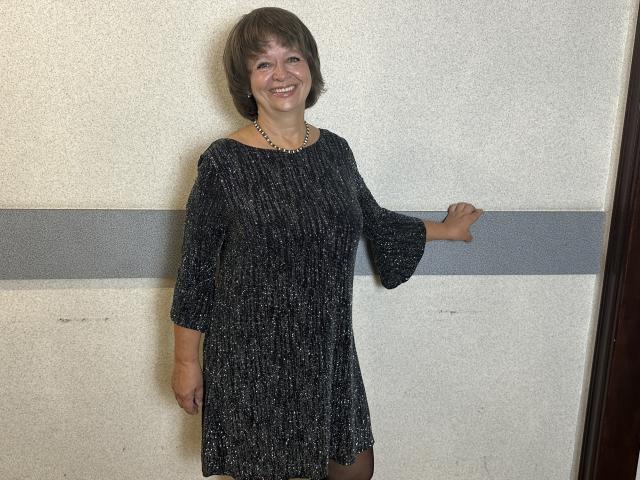 MaltaFora with short brown hair, smiling, wearing a black textured dress with 3/4 sleeves, and a gold necklace, standing against a white wall with a gray stripe.