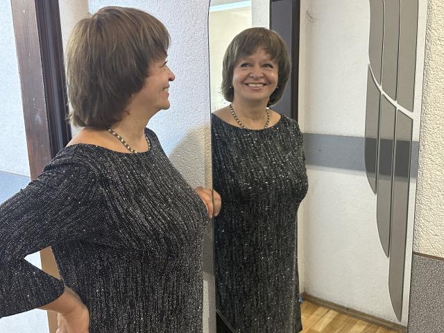 MaltaFora stands in front of a mirror, smiling, wearing a sparkly black dress, her short brown hair, necklace, and hands on her hips. Reflection shows same.