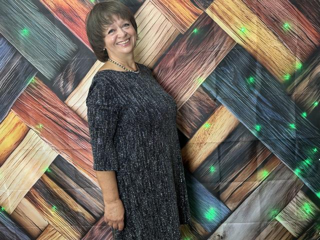 MaltaFora with short brown hair, smiling, wearing a dark, textured dress with a beaded necklace, standing against a colorful, patterned wall with green lights.