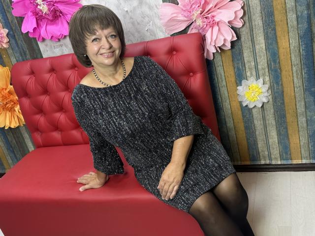 MaltaFora, middle-aged woman with short brown hair, smiling, wearing a black knitted dress, black tights, and a bead necklace, sitting on a red tufted couch.