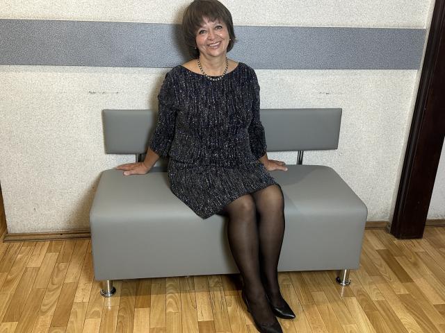 MaltaFora with short brown hair, wearing a black sparkly dress, black pantyhose, and black shoes, sitting on a grey bench, smiling, hands on bench, wooden floor.