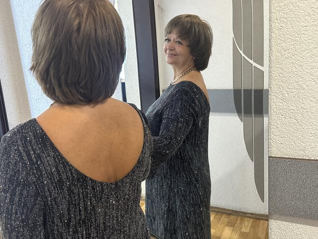 MaltaFora with short brown hair, wearing a sparkly black dress, stands in front of a mirror, smiling at her reflection, showing her back and bare upper back.